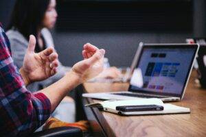 The featured image shows a person's hands positioned in a way that suggests they are explaining something or offering an idea. They are sat at a meeting table with other people and they have a laptop in front of them. Photo by Headway on Unsplash