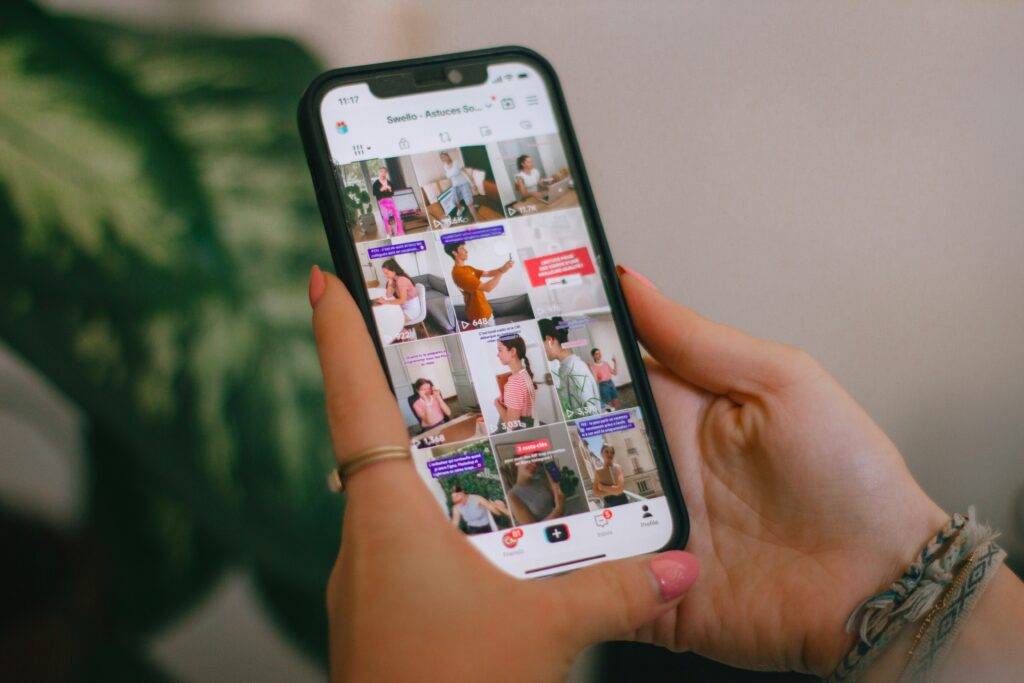 The image is zoomed into a person's hands. They are wearing pink nail polish, fabric bracelets and a ring on their index finger. They are holding a mobile phone which shows a TikTok grid. The background is blurred but there is a large leaf from a plant. Photo by Swello on Unsplash